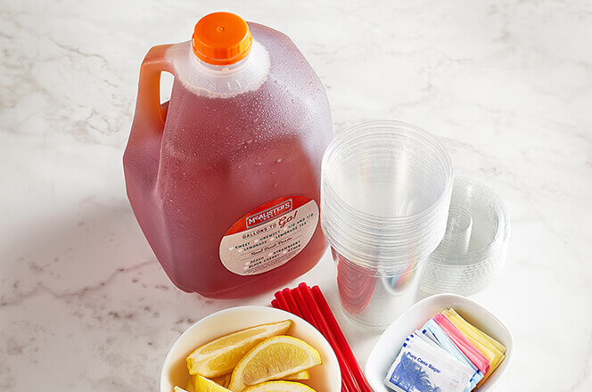 A gallon of McAlister's famous tea, with plastics cups, sliced lemon and packets of sugar on the side of it. 