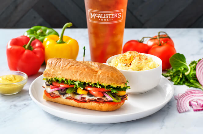 A 6 inch Italian sub sliced in halves, with McALister's famous tea and a side of potatoe salad. Fresh Vegteables in the background. 
