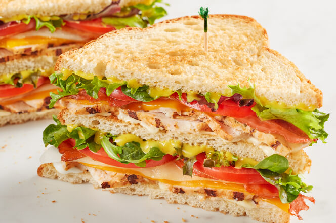 A Grilled Chicken King Club sandwich featuring grilled chicken, bacon, cheddar, Swiss, lettuce, tomato, and honey mustard on wheat bread.