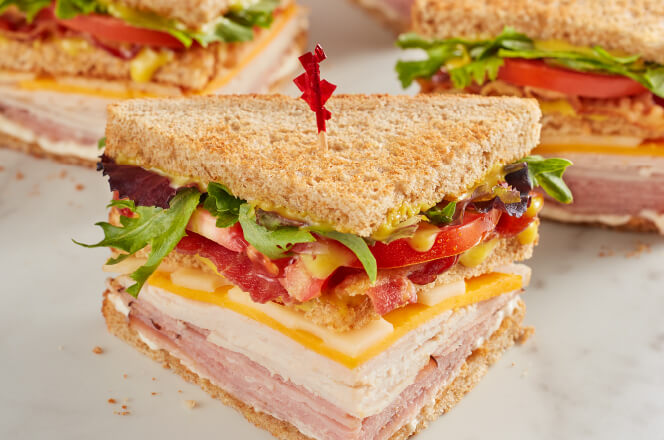 A McAlister's Club sandwich featuring turkey, ham, bacon, cheddar, Swiss, lettuce, tomato, and honey mustard on wheat bread.